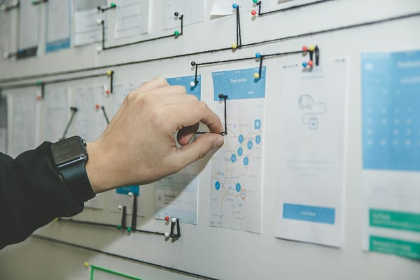 掲示板上の業務フロー資料を指し示す手元。情報整理と業務可視化の取り組みを表しています。