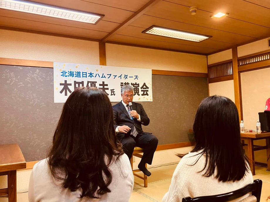 北海道日本ハムファイターズの木田優夫GM代行が、講演会で椅子に座りマイクを持って話している様子を、会場の参加者越しに撮影した写真。