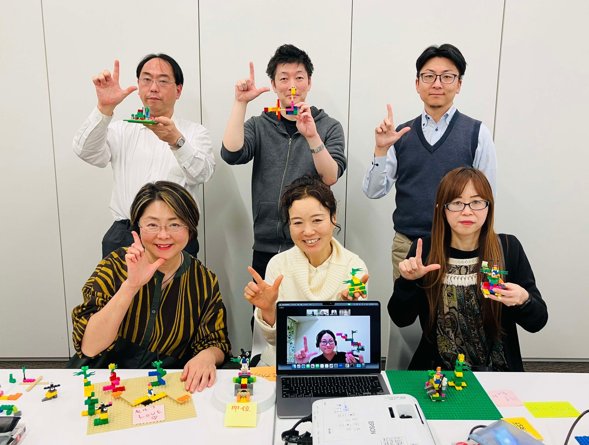 複数の参加者がレゴ®ブロック作品を手に掲げ、「L」を形作るポーズで集合写真。オンライン参加者の姿も画面に映る。